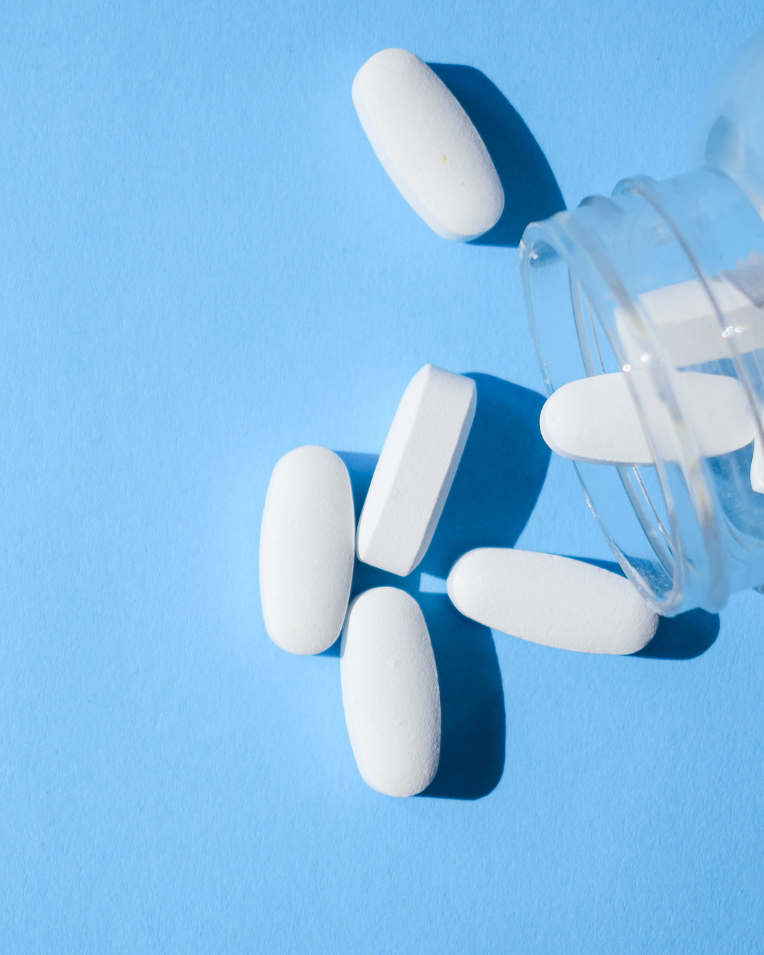 white oxycodin pills resting on a light blue background, having spilled out of a clear pill bottle on it's side and partially out of frame to the right